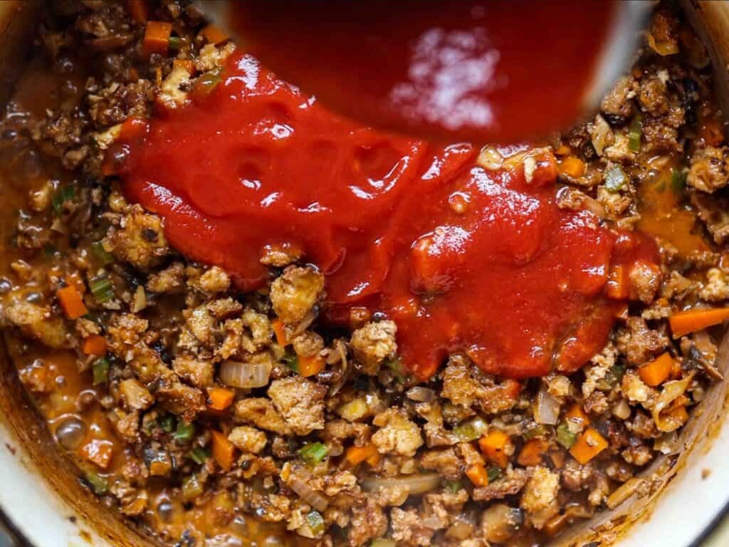 Chopped vegetables, tofu, and vegan sausage are being cooked together in a pot, with tomato sauce being poured over the mixture.